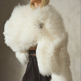 A model wears a fluffy, cropped ivory shearling jacket with black trousers and a black baseball cap, standing before a textured gray concrete wall.