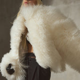 A model in a voluminous, fluffy cropped ivory shearling jacket, paired with black trousers and a black baseball cap, set against a textured gray concrete wall.