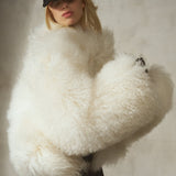 Ivory shearling jacket, cropped and voluminous, styled with black trousers and a black baseball cap on a model against a textured gray concrete wall.