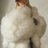 Against a textured gray concrete wall, a model wears a voluminous cropped ivory shearling jacket, black trousers, and a black baseball cap.