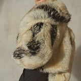 A model wearing a voluminous, shaggy faux fur jacket with a natural beige and black mottled pattern, styled with a black baseball cap and black trousers against a textured gray wall.