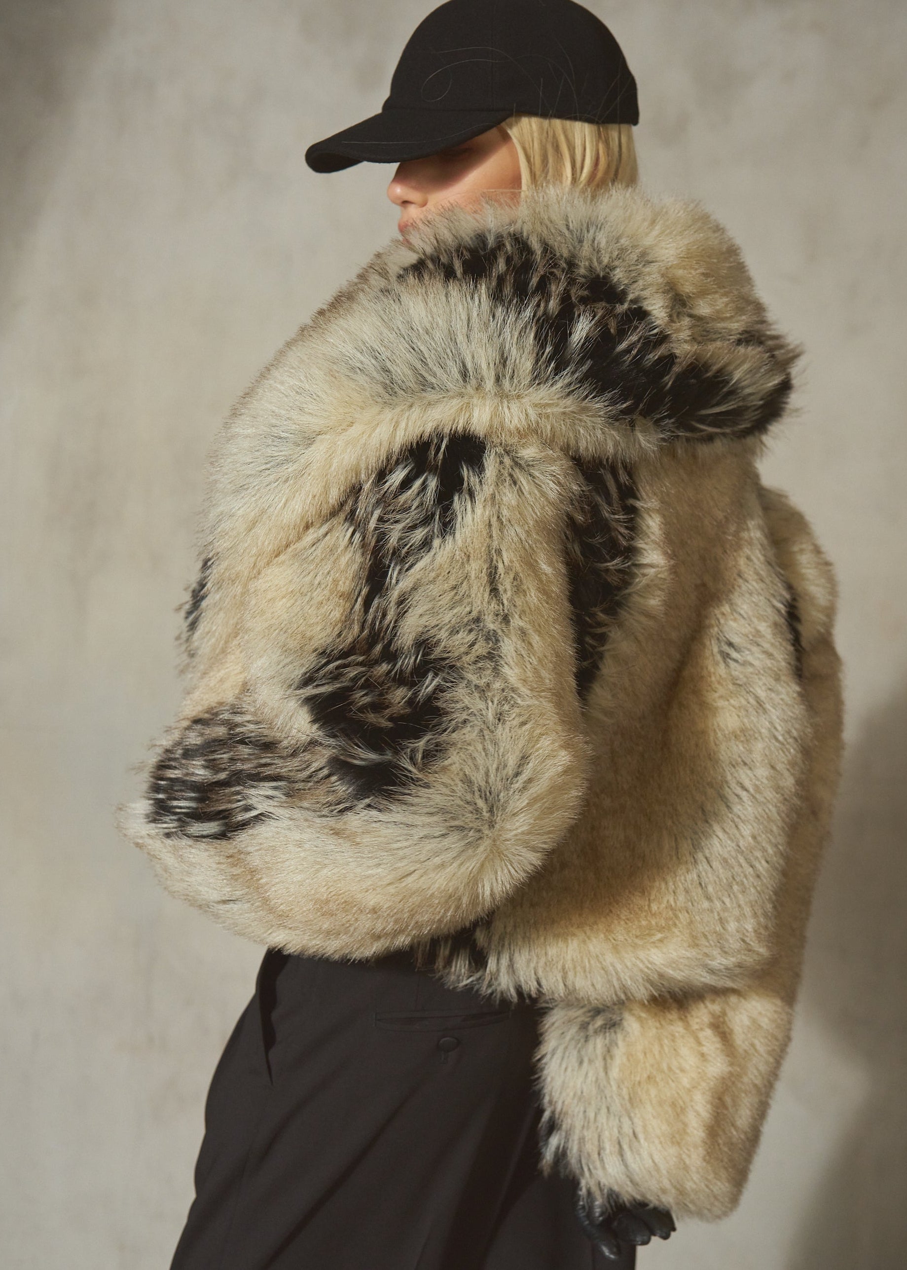 A model wearing a voluminous, shaggy faux fur jacket with a natural beige and black mottled pattern, styled with a black baseball cap and black trousers against a textured gray wall.