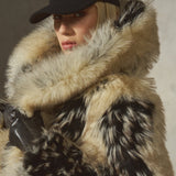 A model wearing a luxurious black and beige mottled faux fur coat with a large matching faux fur collar, a black baseball cap, and black gloves, set against a textured gray wall.