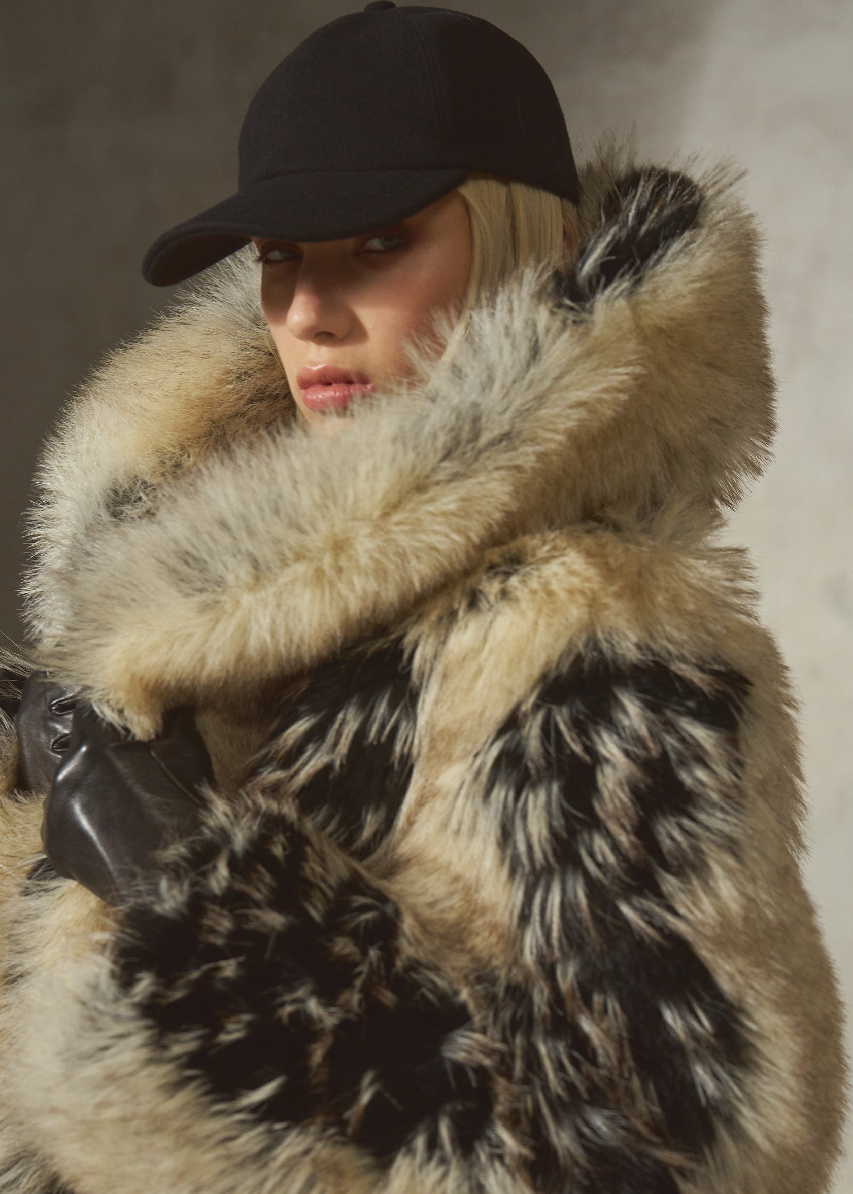 A model wearing a luxurious black and beige mottled faux fur coat with a large matching faux fur collar, a black baseball cap, and black gloves, set against a textured gray wall.
