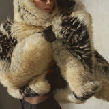 A model wearing a voluminous black and beige faux fur coat with a matching faux fur-lined collar, a black cap, and black pants, posed against a textured gray wall.
