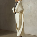 A model in a cashmere-wool blend, cream-colored wrap coat with wide draped sleeves and belted at the front, styled with black leather gloves, a black cap, sunglasses, black tights, and black high heels, set against a textured concrete wall.