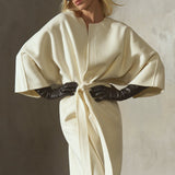 A model styled in a cream-colored, cashmere-wool blend wrap coat with wide draped sleeves and a front belt, a black cap, sunglasses, and black leather gloves,  photographed against a textured concrete wall.