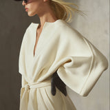 Dressed in a cashmere-wool blend, cream-colored wrap coat with wide draped sleeves and a front belt, a model wears a black cap, sunglasses, and black leather gloves, against a textured concrete wall.