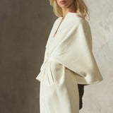 A model wearing a cashmere-wool blend, cream-colored wrap coat with wide draped sleeves, belted at the front, styled with a black cap, sunglasses and black leather gloves, standing against a textured concrete wall.