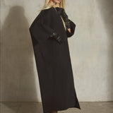 A black cashmere-wool blend wrap coat with wide draped sleeves, is styled with a black cap, black leather gloves, black tights, and black high heels on a model against a textured concrete wall.