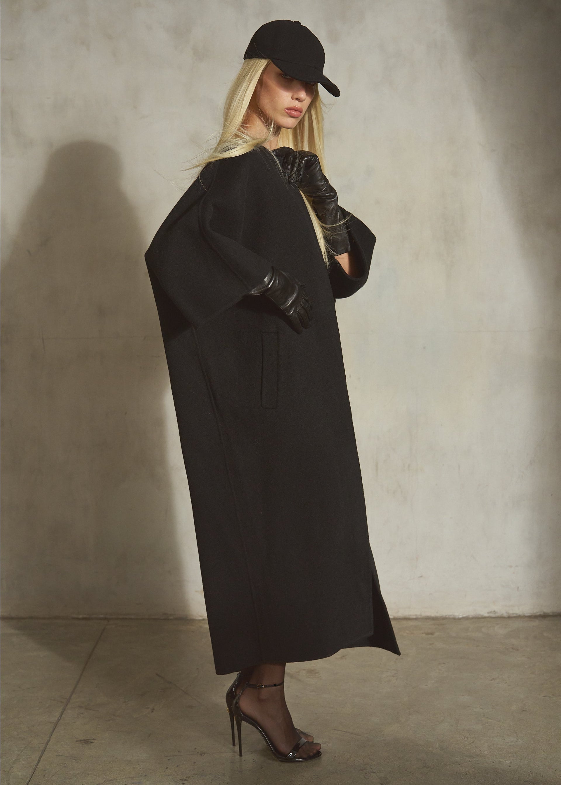 A black cashmere-wool blend wrap coat with wide draped sleeves, is styled with a black cap, black leather gloves, black tights, and black high heels on a model against a textured concrete wall.