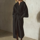 Against a textured concrete wall, a model wears a black cashmere-wool blend wrap coat with wide draped sleeves, belted in the front, styled with a black cap, black leather gloves, black tights, and black high heels.