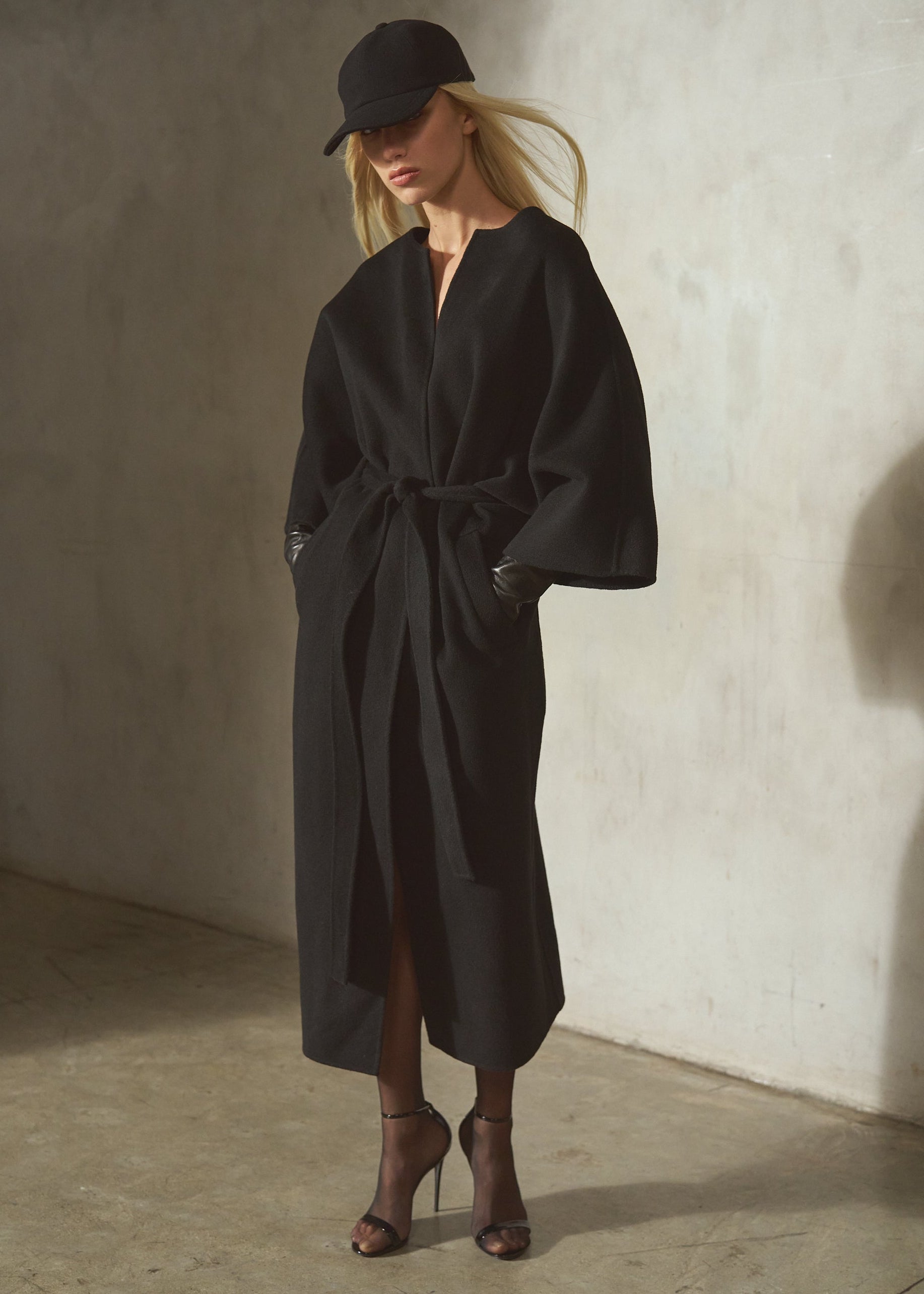 Against a textured concrete wall, a model wears a black cashmere-wool blend wrap coat with wide draped sleeves, belted in the front, styled with a black cap, black leather gloves, black tights, and black high heels.