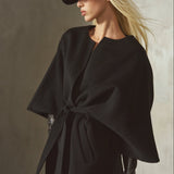 A model wears a black cashmere-wool blend wrap coat with wide draped sleeves, belted at the front, completed by a black cap and black leather gloves, against a textured concrete wall.