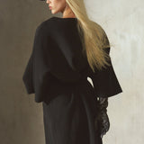 Dressed in a black cashmere-wool blend wrap coat with wide draped sleeves and a front belt, a model is styled with a black cap, and black leather gloves, against a textured concrete wall.