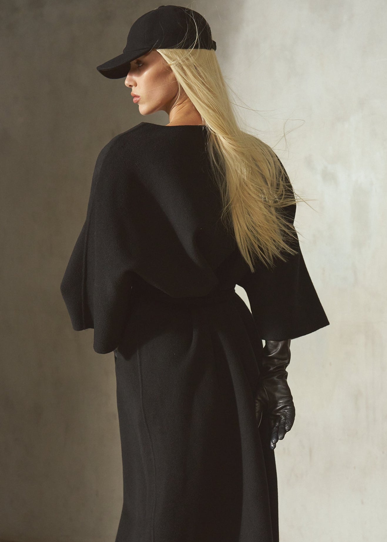 Dressed in a black cashmere-wool blend wrap coat with wide draped sleeves and a front belt, a model is styled with a black cap, and black leather gloves, against a textured concrete wall.