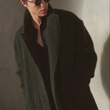 A man wearing a dark green textured wool-blend coat over a black shirt and black sunglasses, standing against a textured gray wall.