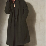 A model wearing a long, dark olive green wool overcoat, black trousers, a black shirt, and black gloves, standing against a textured concrete wall.