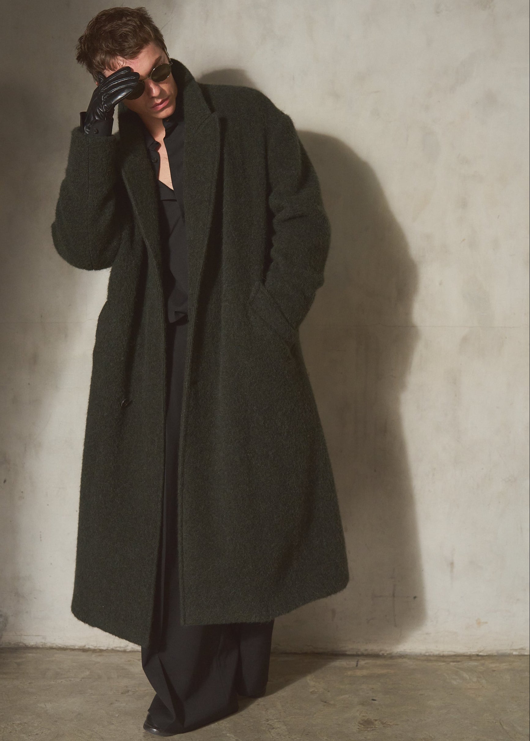 A model wearing a long, dark olive green wool overcoat, black trousers, a black shirt, and black gloves, standing against a textured concrete wall.