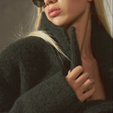 A blonde woman wearing dark brown cat-eye sunglasses and a dark green fuzzy coat, captured in a moody, warm-toned studio portrait.