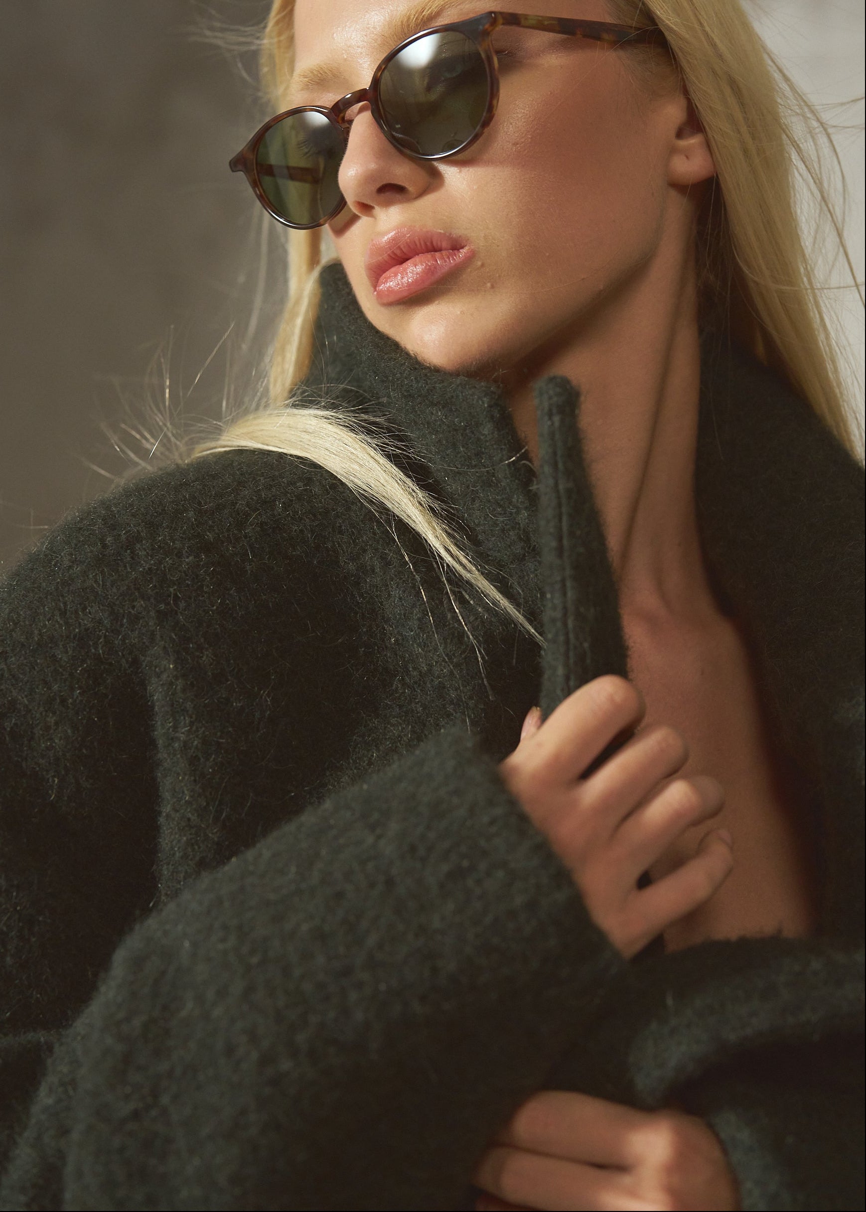 A blonde woman wearing dark brown cat-eye sunglasses and a dark green fuzzy coat, captured in a moody, warm-toned studio portrait.