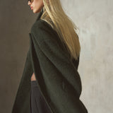 A blonde woman in side profile wearing a dark olive green oversized coat and black pants, standing against a textured gray wall.