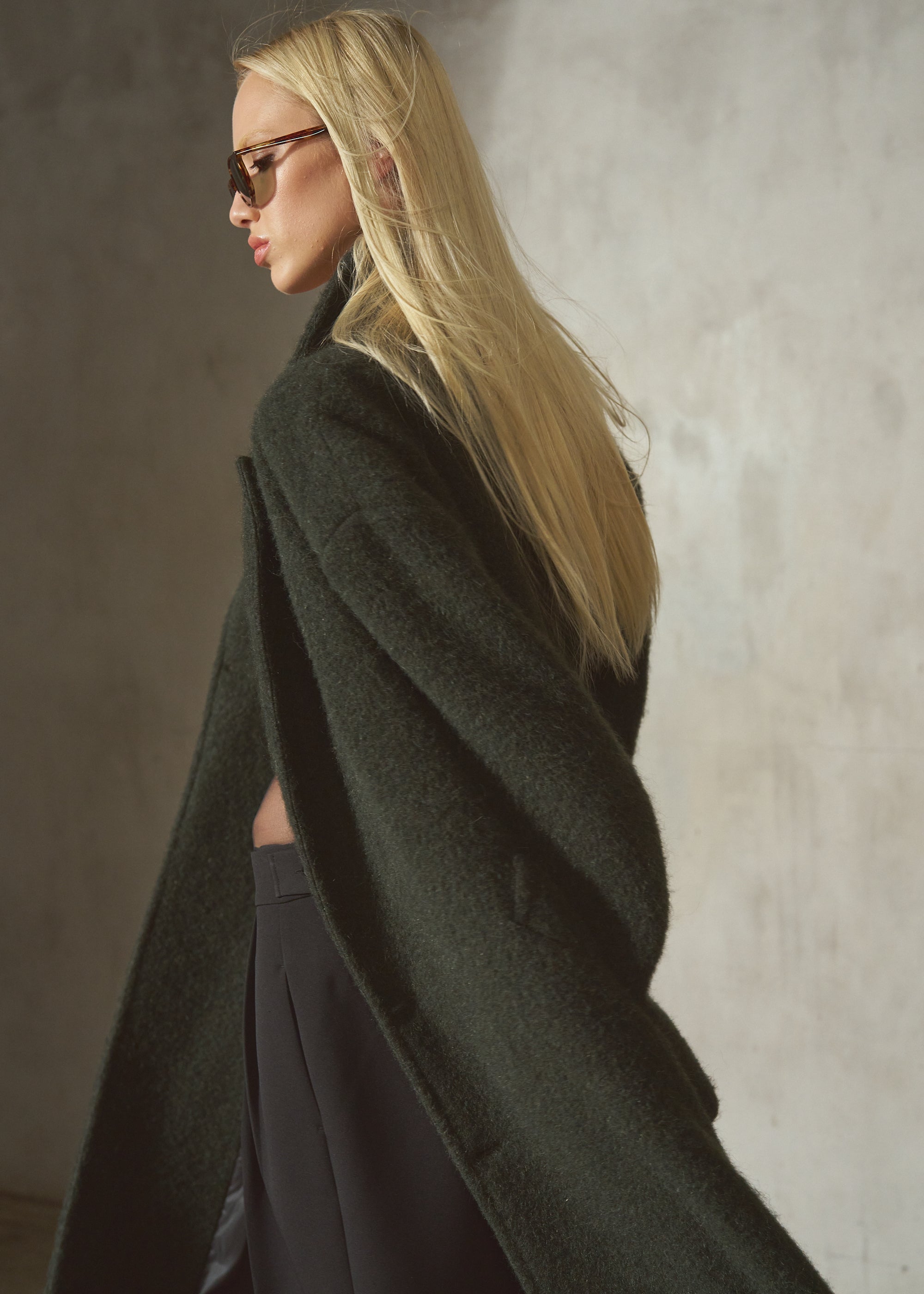 A blonde woman in side profile wearing a dark olive green oversized coat and black pants, standing against a textured gray wall.