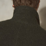 Close-up back view of a person wearing a dark olive green wool coat with a high collar, showing the texture of the fabric and hair against a neutral background.