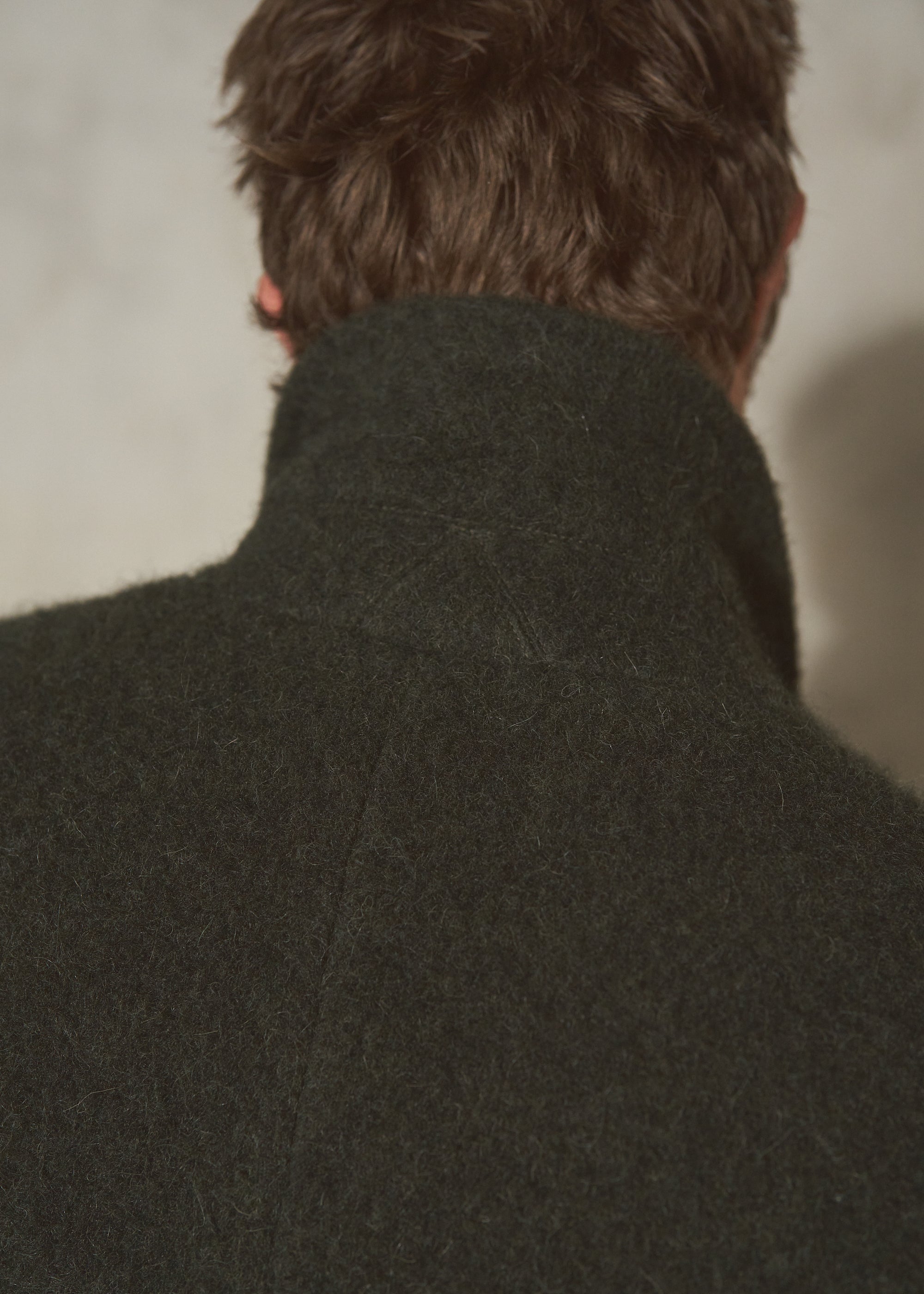 Close-up back view of a person wearing a dark olive green wool coat with a high collar, showing the texture of the fabric and hair against a neutral background.