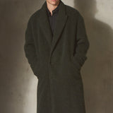 A man wearing a long dark olive green wool-blend coat over a black shirt and wide-leg black trousers, standing against a textured concrete wall with a strong shadow.