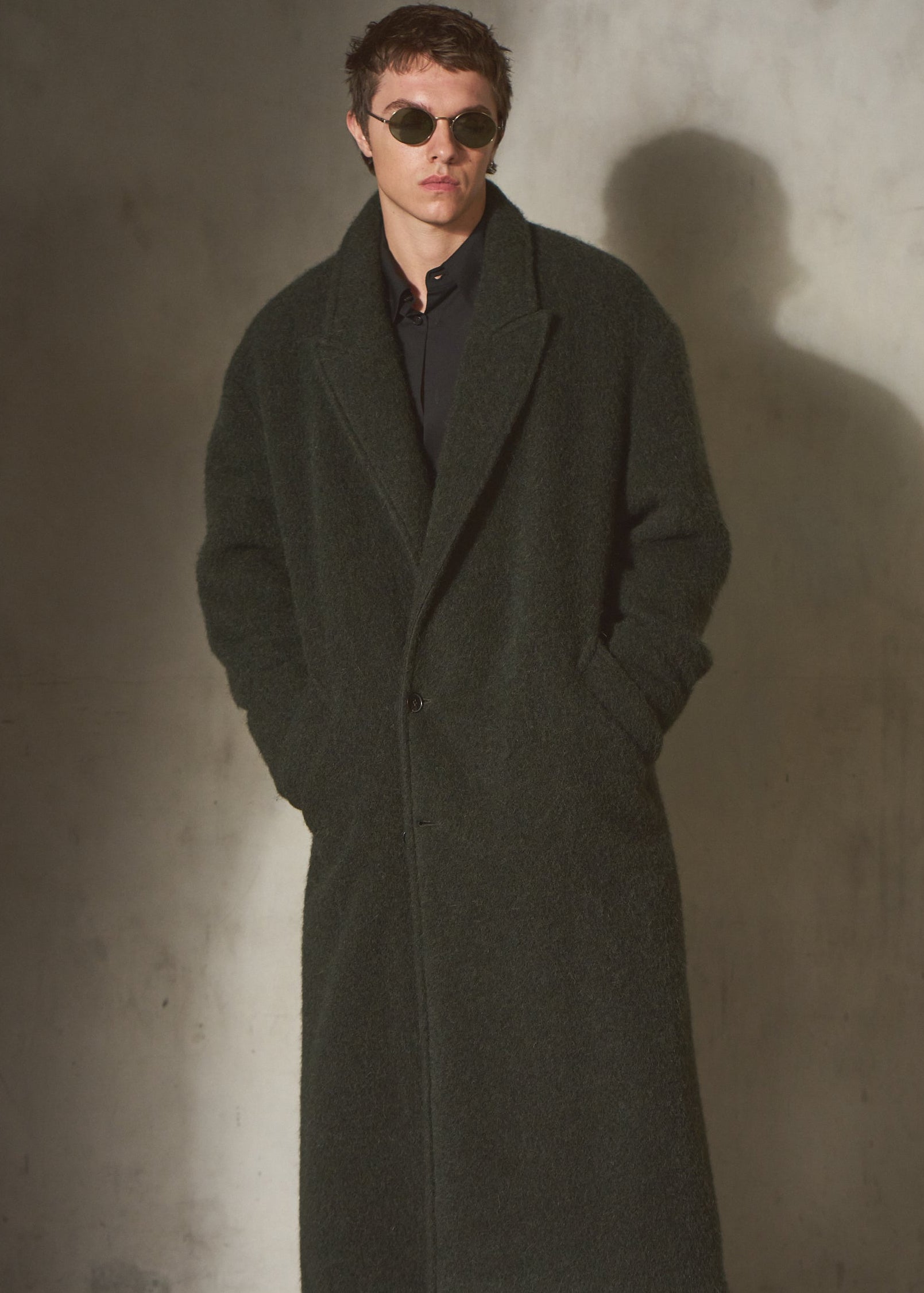 A man wearing a long dark olive green wool-blend coat over a black shirt and wide-leg black trousers, standing against a textured concrete wall with a strong shadow.