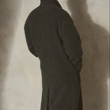 A man viewed from behind wearing a long dark olive green wool-blend coat over dark trousers, standing against a textured concrete wall with a strong shadow.