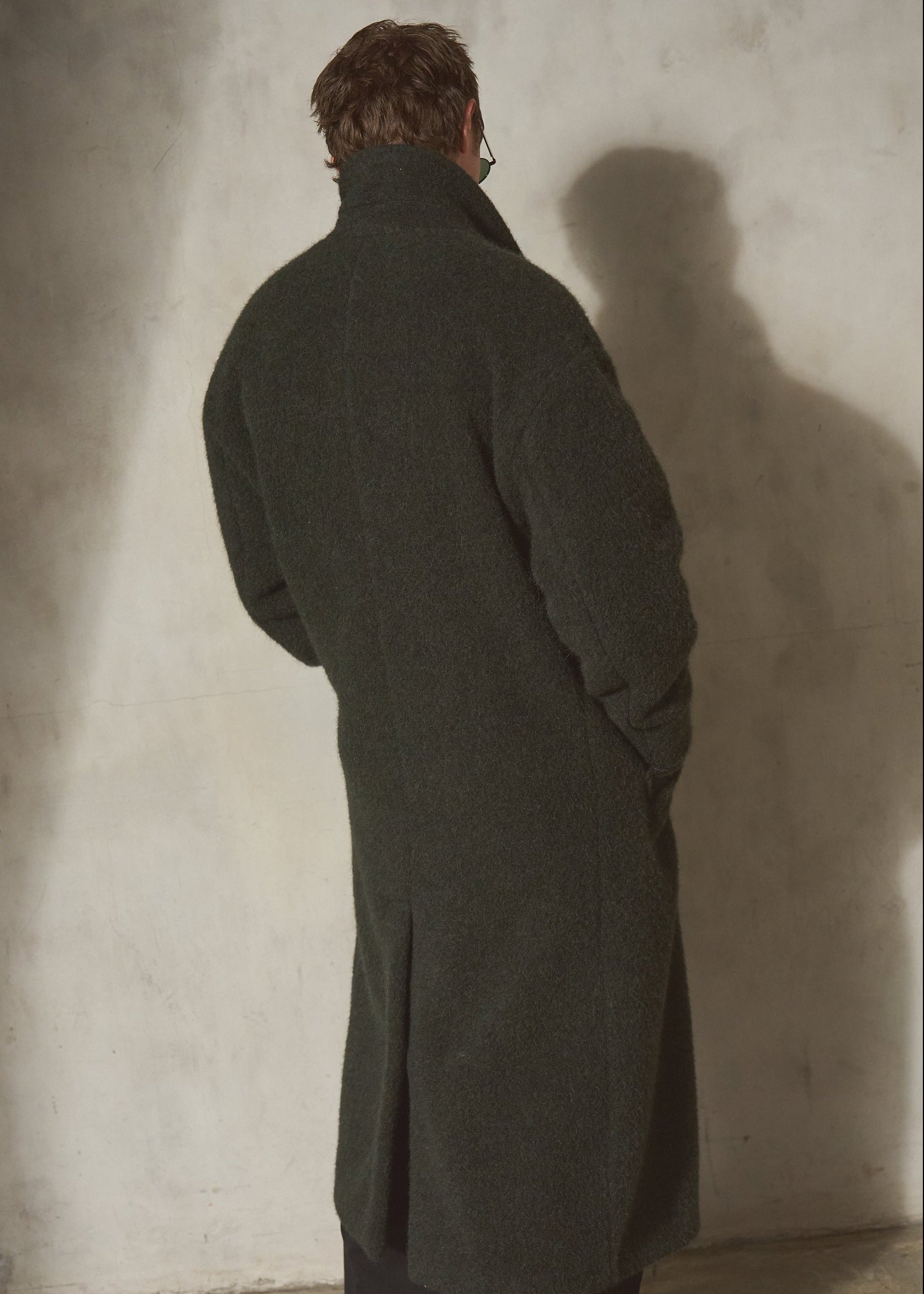 A man viewed from behind wearing a long dark olive green wool-blend coat over dark trousers, standing against a textured concrete wall with a strong shadow.
