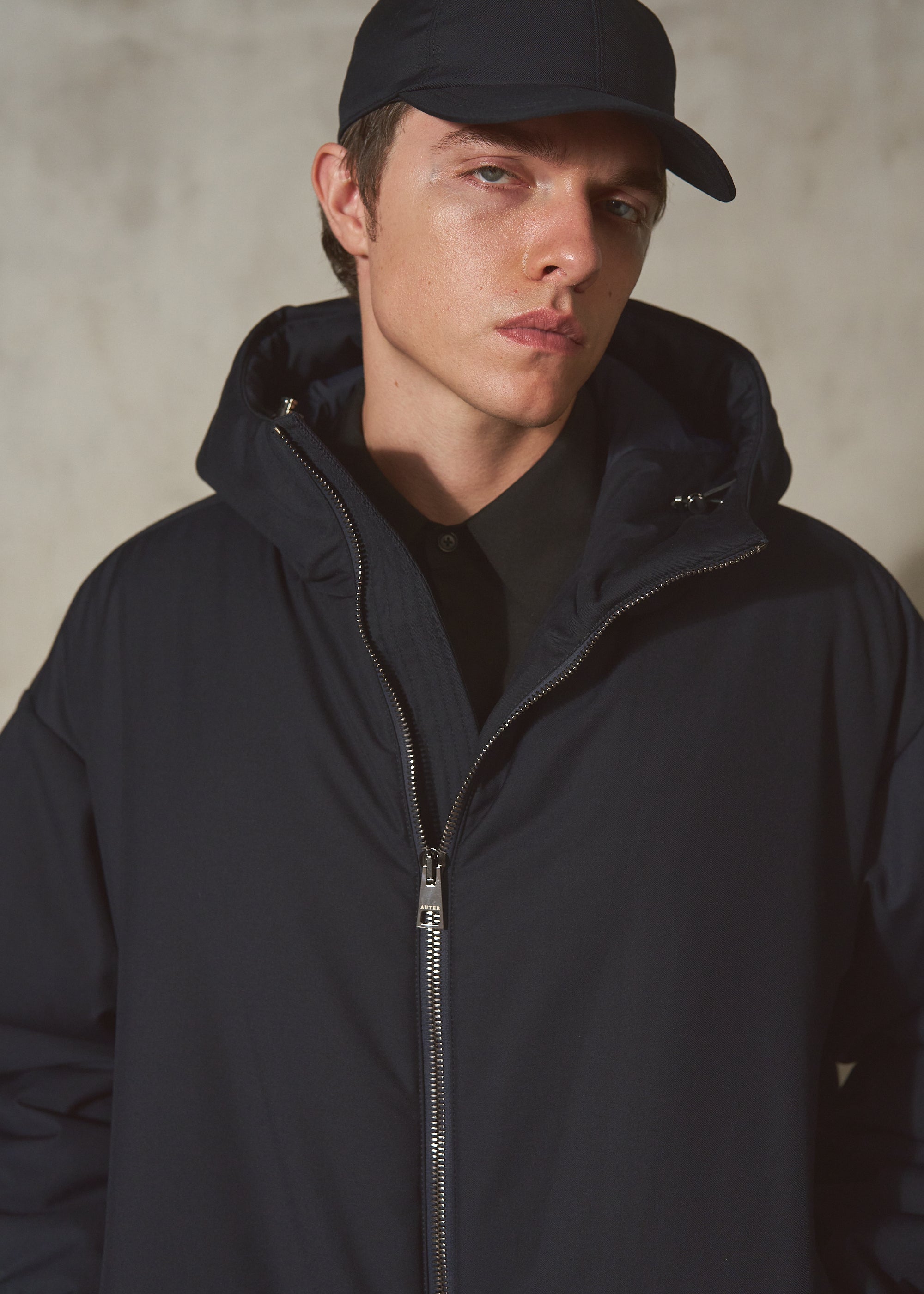 A man wearing a dark navy hooded zip-up jacket and a matching dark baseball cap, photographed against a textured concrete wall.