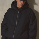 Model wearing a navy hooded zip-up jacket, navy cap, and black gloves, standing against a textured concrete wall with dramatic side lighting.