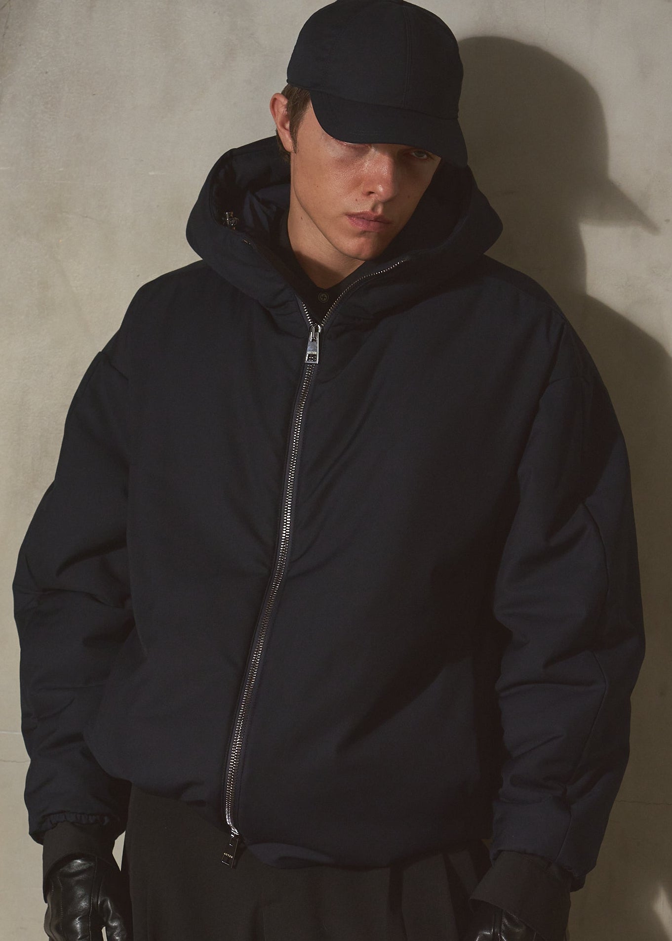 Model wearing a navy hooded zip-up jacket, navy cap, and black gloves, standing against a textured concrete wall with dramatic side lighting.