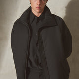 A model wearing a black cap, black oversized jacket, black shirt, and black trousers, standing against a textured concrete wall with a strong shadow cast to the side.