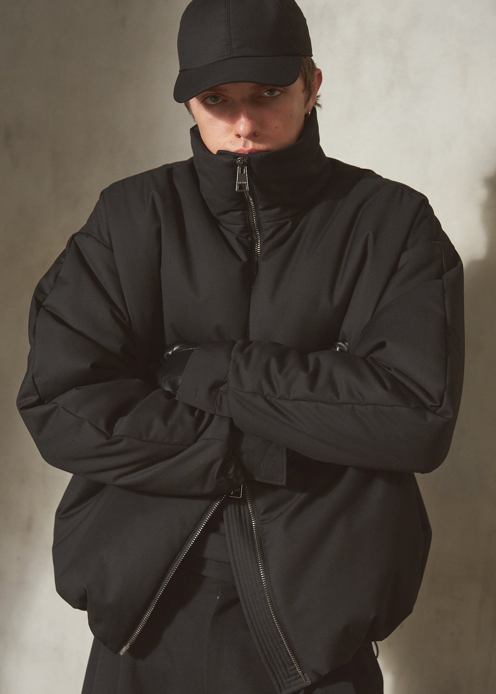 A male model wearing a black puffer jacket with a high collar, black pants, and a black baseball cap, standing against a textured concrete wall with arms crossed.