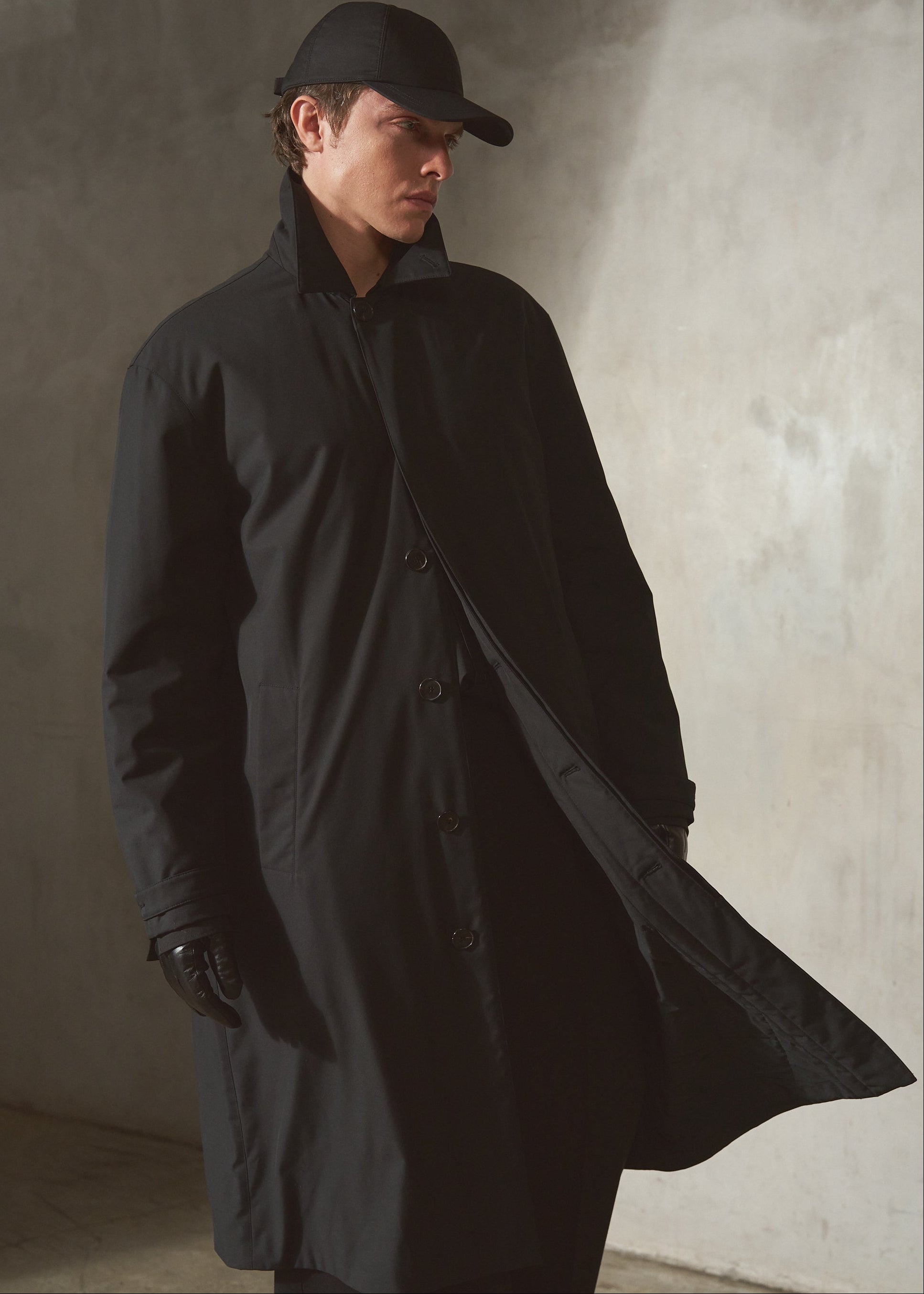 A man wearing a long black mac coat, with a matching black cap, and black gloves, standing against a textured concrete wall.