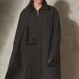 A male model wearing a long black quilted trench coat, black pants, a black cap, and black gloves, standing against a textured concrete wall with dramatic side lighting.