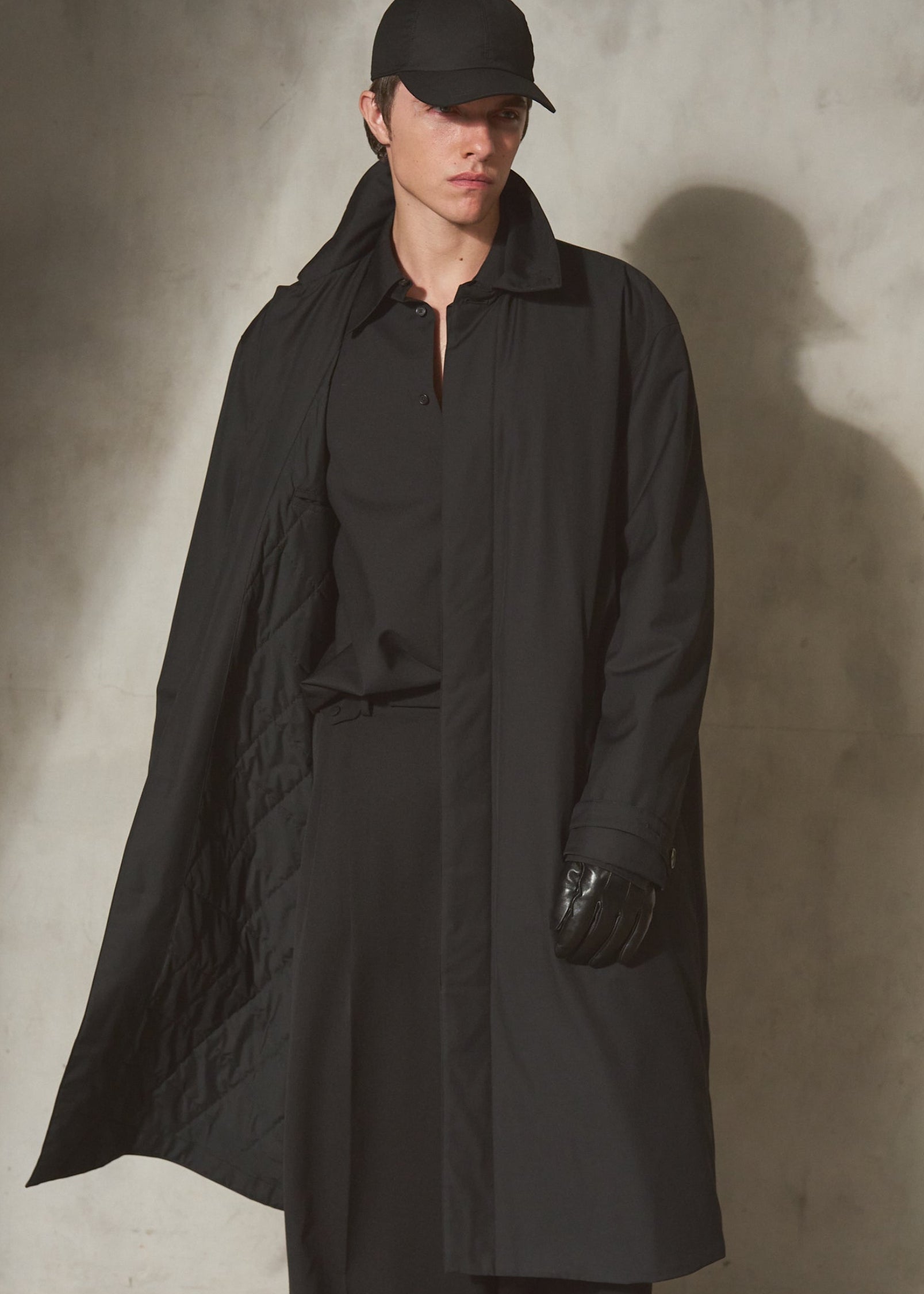 A male model wearing a long black quilted trench coat, black pants, a black cap, and black gloves, standing against a textured concrete wall with dramatic side lighting.