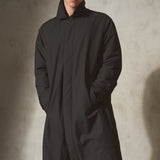 A model wearing a long black mac coat, black pants, and a matching black cap stands against a textured concrete wall with dramatic side lighting.