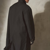 A model wearing a long black trench coat, black baseball cap, and black gloves, standing in profile against a textured gray concrete wall.