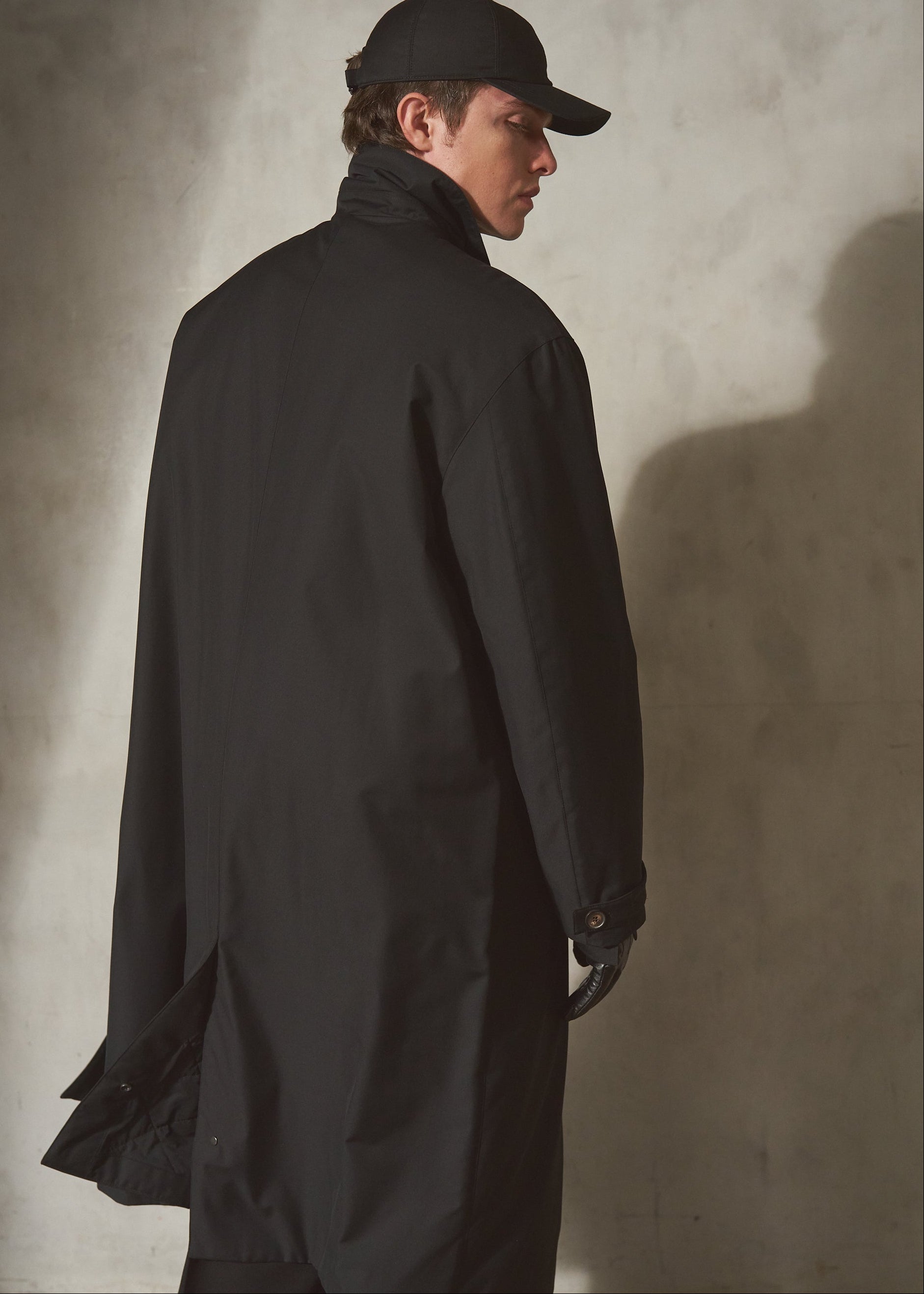 A model wearing a long black trench coat, black baseball cap, and black gloves, standing in profile against a textured gray concrete wall.