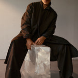 Model wearing a dark black long mac coat sitting on a transparent ice block with a plain background.