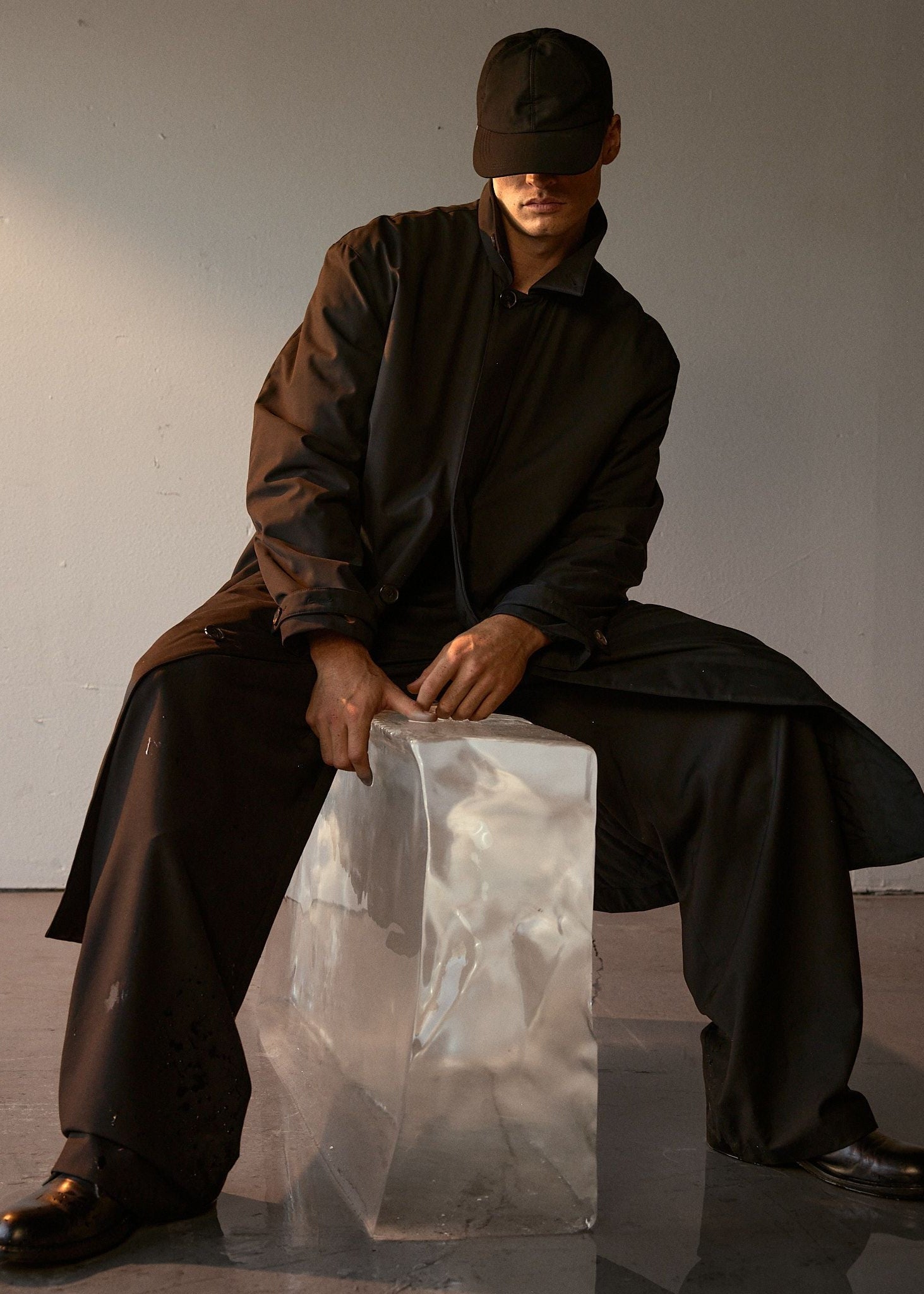 Model wearing a dark black long mac coat sitting on a transparent ice block with a plain background.
