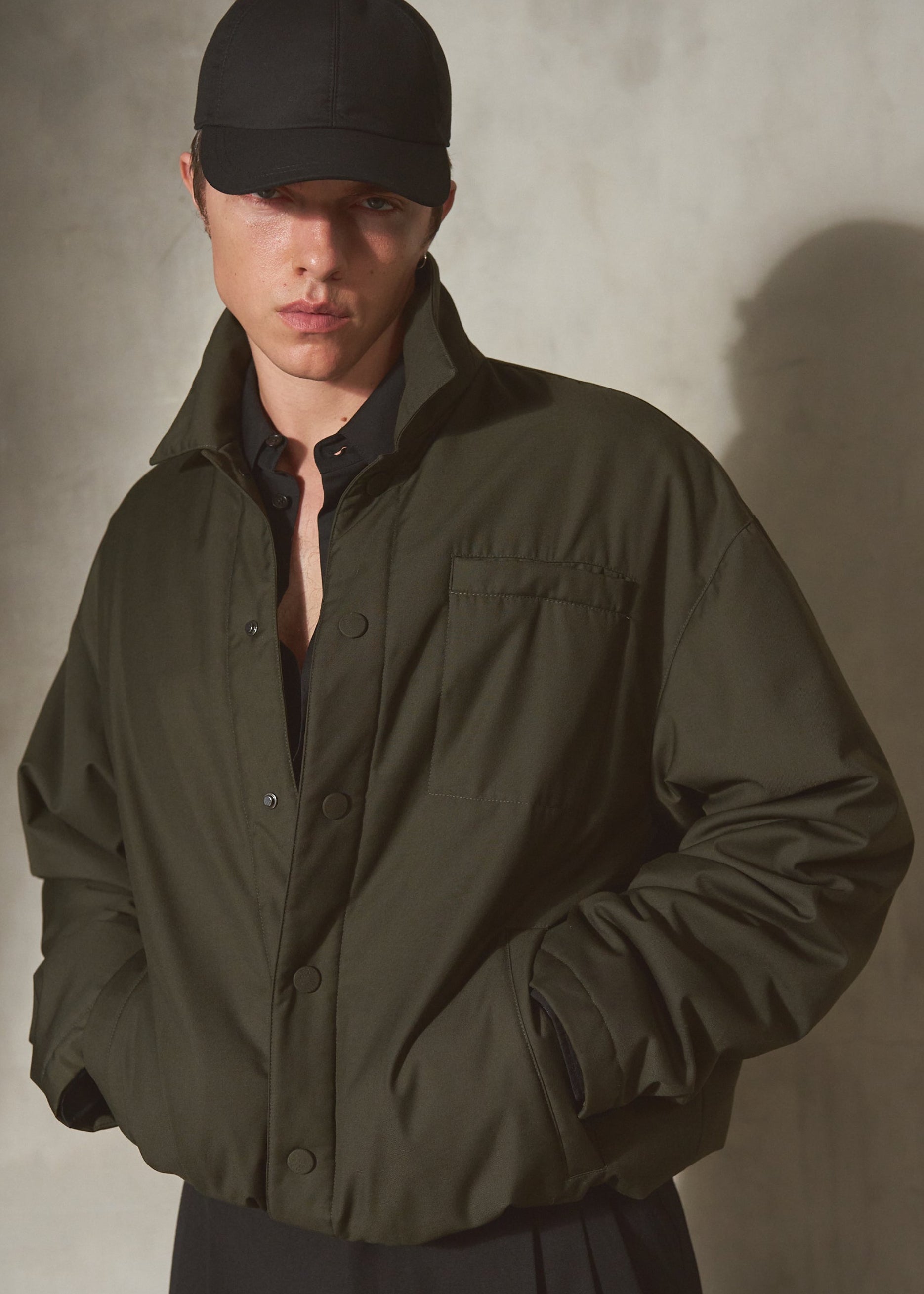 A model wearing an oversized dark olive green utility jacket, a black baseball cap, and a black shirt, standing against a textured gray wall with dramatic lighting.
