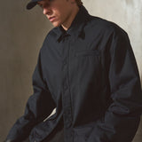 A man wearing a dark navy blue button-up shirt jacket, navy baseball cap, black pants, and black leather gloves, leaning against a textured concrete wall.