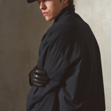 A man wearing a navy baseball cap, navy jacket, and black gloves, sitting against a textured concrete wall in warm, directional light.