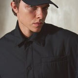 A close-up of a man wearing a navy baseball cap and a navy button-up shirt jacket against a textured beige wall.